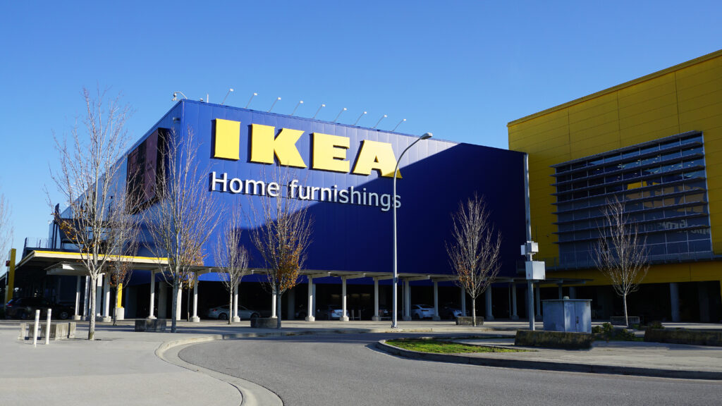 The image showcases an IKEA store with its iconic blue and yellow exterior, perfectly designed to attract consumer attention using marketing psychology. The facade displays the large IKEA logo and the words "Home furnishings," framed by a road and leafless trees under a clear blue sky.