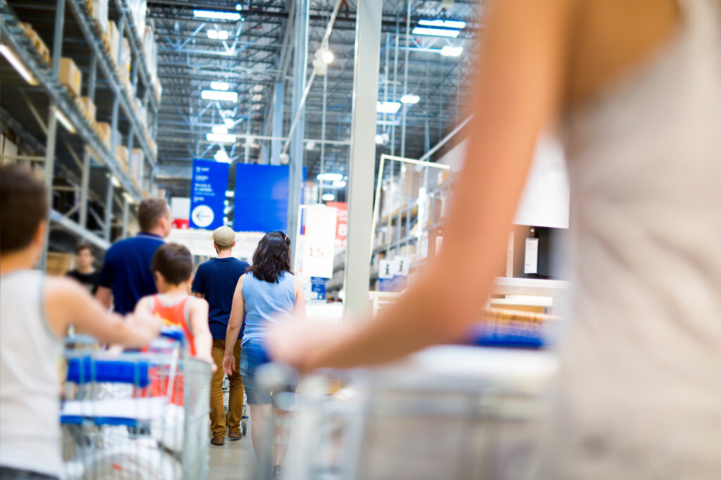 Shoppers navigate a spacious warehouse-style store with high shelves and overhead lighting, exemplifying consumer behavior. Customers push shopping carts, browsing aisles of diverse products in a busy yet casual atmosphere.