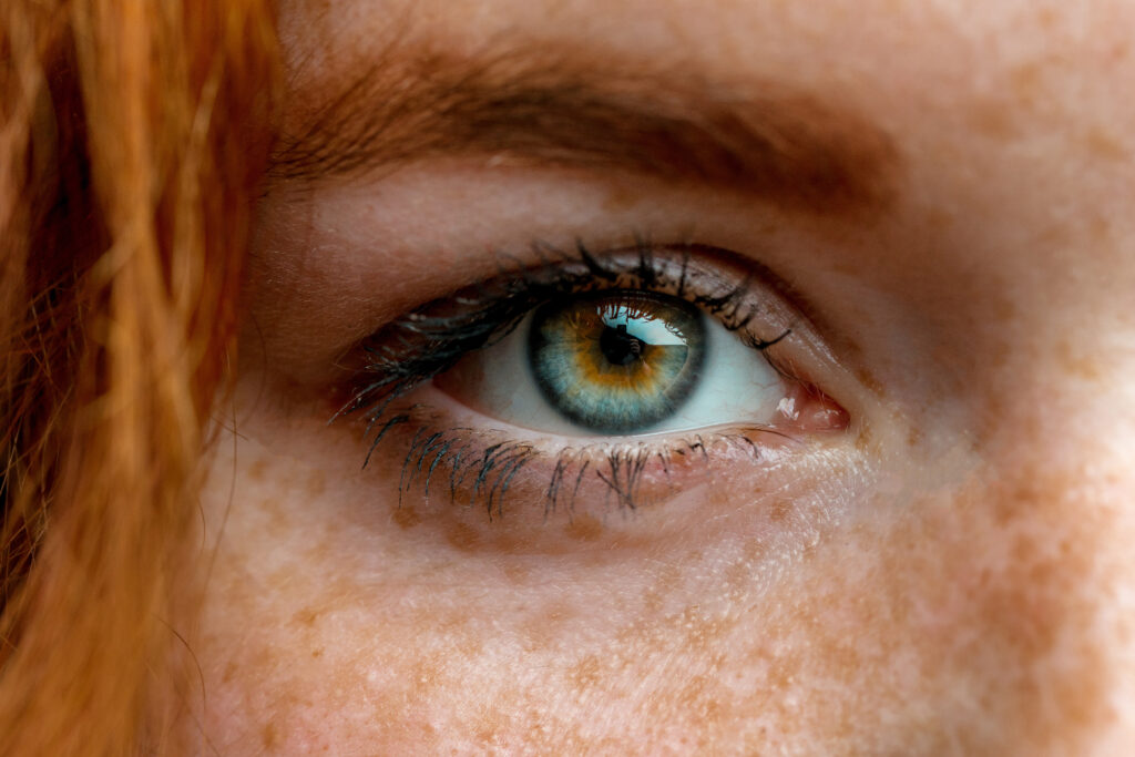 Close-up of a person's eye with green and brown hues, prominent eyelashes, and freckles on the skin. The eyebrow and a hint of red hair are visible, creating a captivating visual akin to the intriguing insights found in behavioral science.
