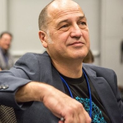 A person with short hair is seated, wearing a dark blazer over a black shirt with a blue geometric design. The person has a thoughtful expression and appears to be in a meeting or conference setting.