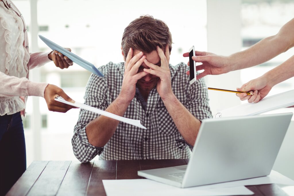 A man sits at a table with his head in his hands, surrounded by outstretched arms holding paperwork, a smartphone, and a pencil. An open laptop suggests an overwhelming work environment influenced by the principles of behavioral economics.