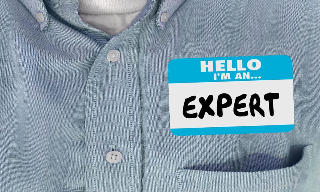 A person wearing a light blue button-up shirt with a name tag on the chest pocket. The name tag has a blue border and reads, "Hello, I'm an... EXPERT" in black, bold letters.