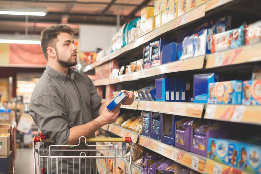 A man with a beard, wearing a gray shirt, is standing in a grocery store aisle holding a blue box, inspecting it. A shopping cart is in front of him, and shelves stocked with various packaged goods are in the background.