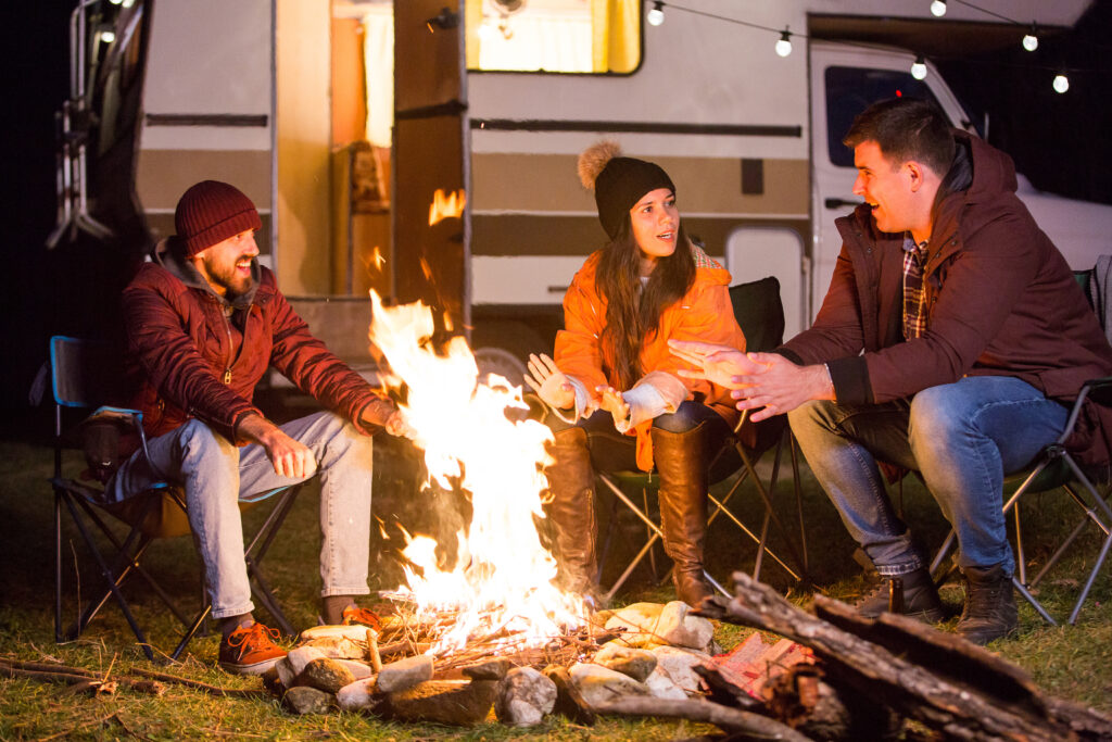 Three people sit around a campfire at night, engaging in conversation about consumer behavior. They are dressed warmly in jackets, hats, and gloves. A camper van, decorated with string lights, is visible in the background, adding a cozy touch to their behavioral science discussions.