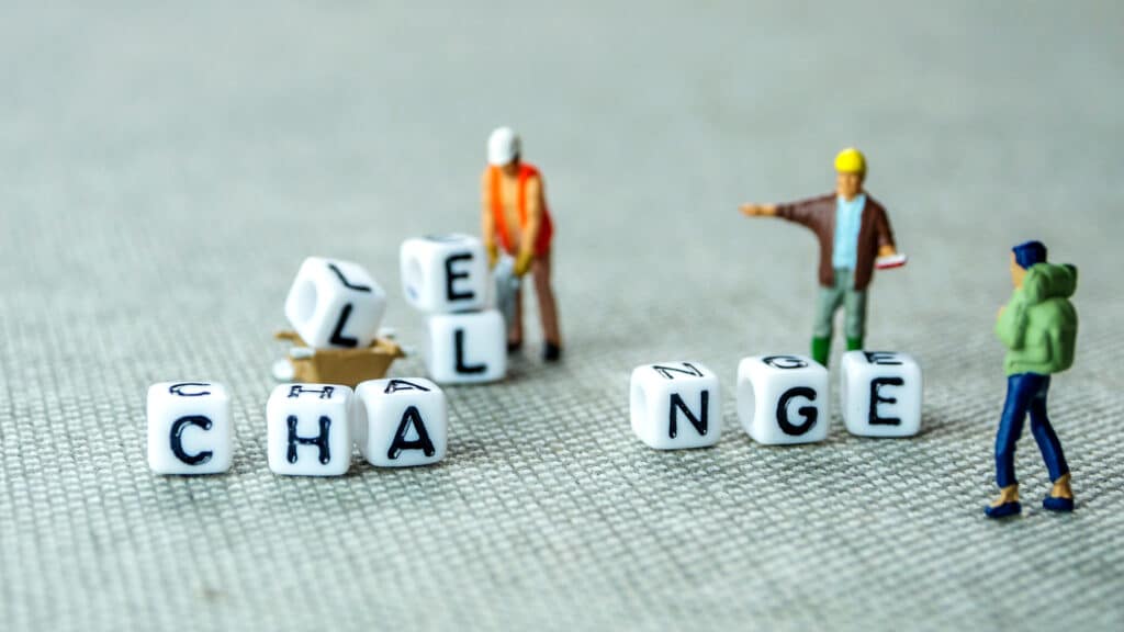 Miniature figures in construction attire interact around letter blocks on a gray surface. The blocks spell "CHANGE," with some letters toppled, suggesting the word "CHALLENGE.