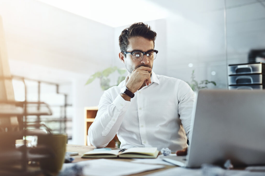 A man in a white shirt sits at a wooden desk, contemplating consumer behavior as he gazes at his laptop. Wearing glasses, he rests his hand on his chin. An open notebook and a cup are on the desk, with sunlight streaming in from the window, setting the perfect scene for marketing insights.