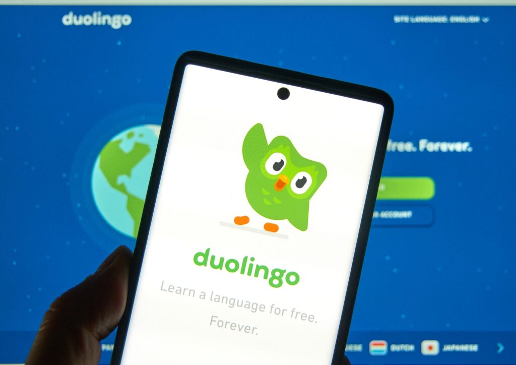 A smartphone displaying the Duolingo app with its green owl mascot is held up against a computer screen showing Duolingo's website homepage. The backdrop features a blue globe graphic, and text promoting language learning.