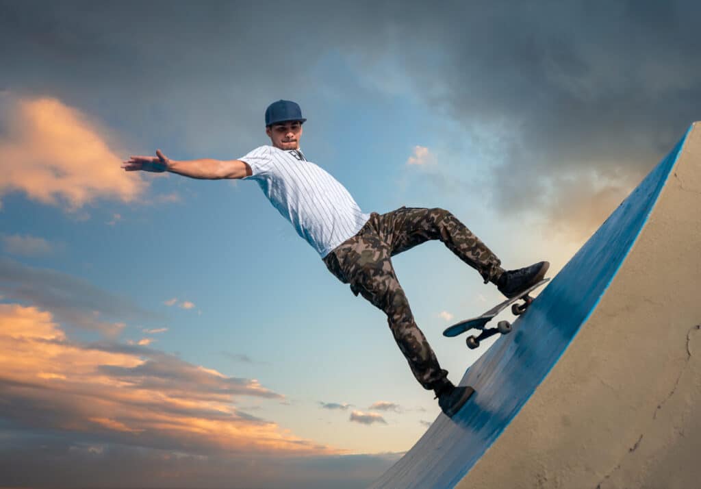 A man wearing a blue cap, striped shirt, and camouflage pants skateboards on a blue ramp against a dramatic sky with clouds and sunset hues. His arms are outstretched for balance.