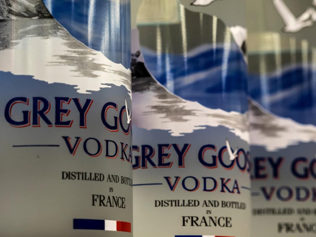 Close-up of several Grey Goose vodka bottles, showing the label which reads "Distilled and Bottled in France" with the French flag at the bottom. The label features a blue and white design with a flying goose.