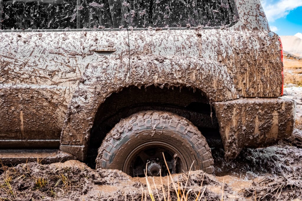 A muddy SUV stuck in thick mud, with one wheel partially submerged, indicating a challenging off-road situation. The vehicle's side is covered in splattered mud, showcasing an adventurous drive through rugged terrain.