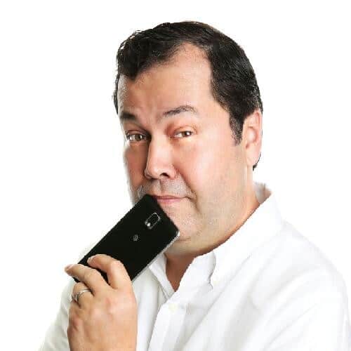 A man in a white shirt holds a black smartphone to his chin while looking directly at the camera with a thoughtful expression. The background is plain white.