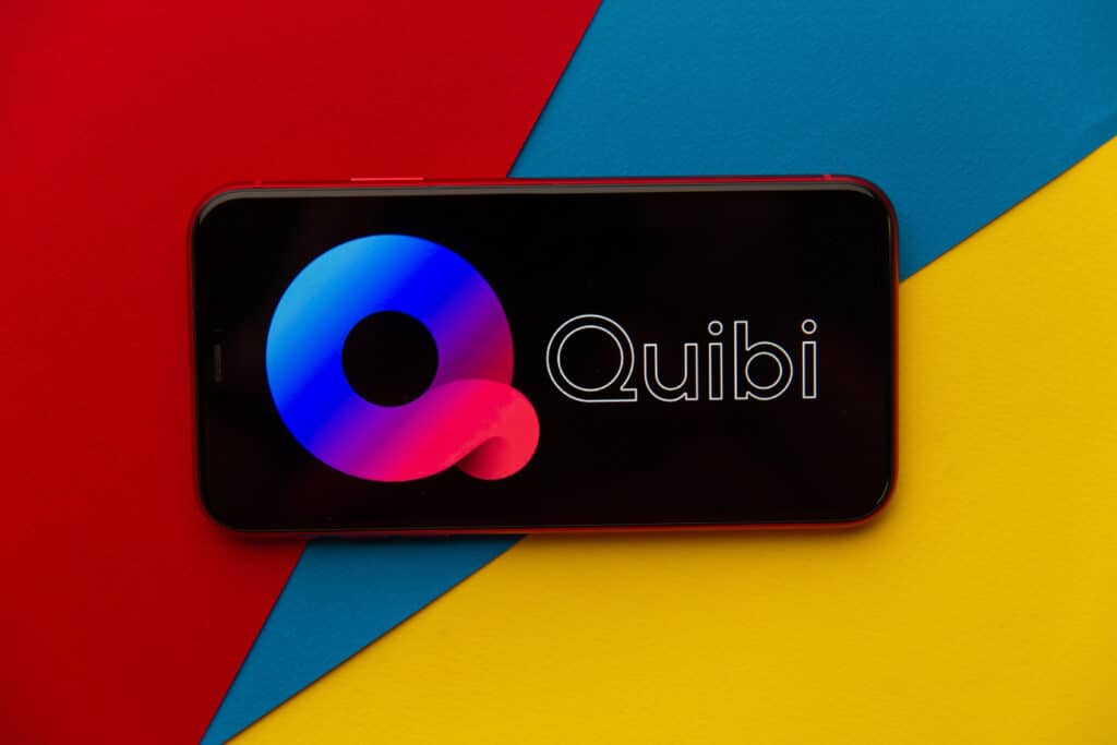 A smartphone displaying the Quibi logo on its screen rests on a vibrant background of red, blue, and yellow diagonal sections, subtly tapping into marketing psychology to capture consumer behavior effectively.