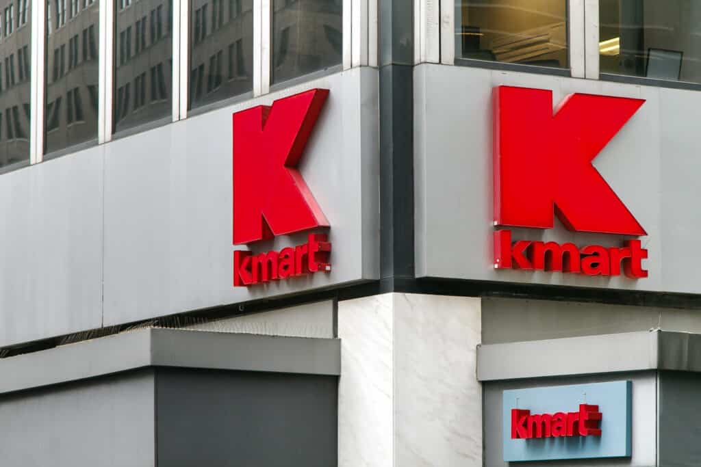 Close-up of a building corner featuring large, red "Kmart" logos on gray panels, designed with an understanding of consumer behavior. The prominent letter "K" is above the smaller word "kmart". The building has reflective windows and an urban city backdrop.