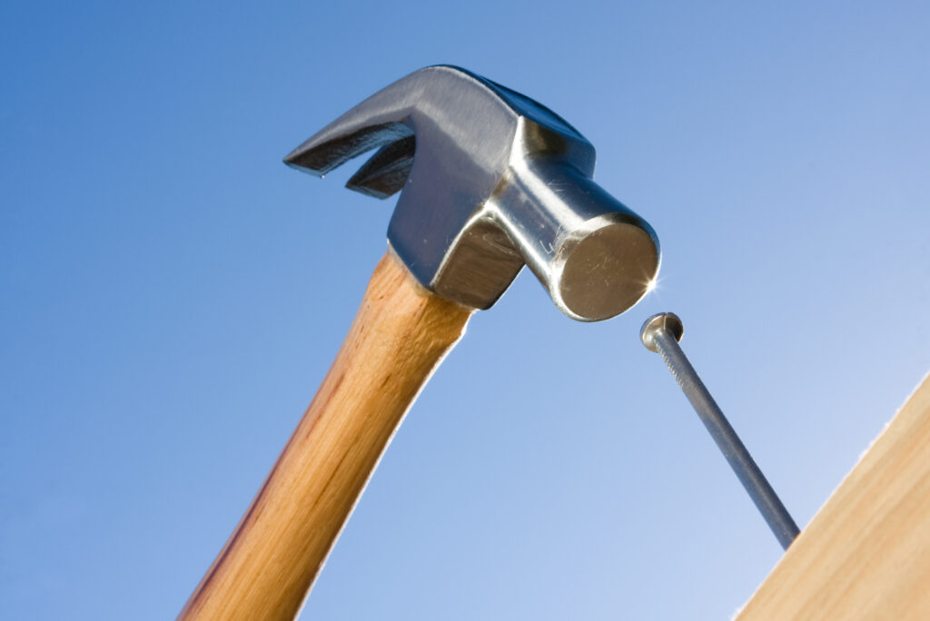 A hammer with a wooden handle is poised above a nail, ready to strike, illustrating the precision often employed in marketing psychology. The scene is set against a clear blue sky.