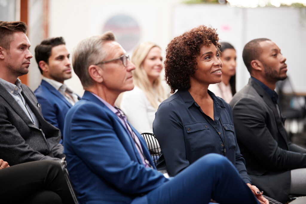 A diverse group of people seated attentively in a professional setting, focused on something ahead, likely discussing marketing psychology. The attendees are dressed in business attire, suggesting a conference or seminar. The atmosphere appears engaged and positive.