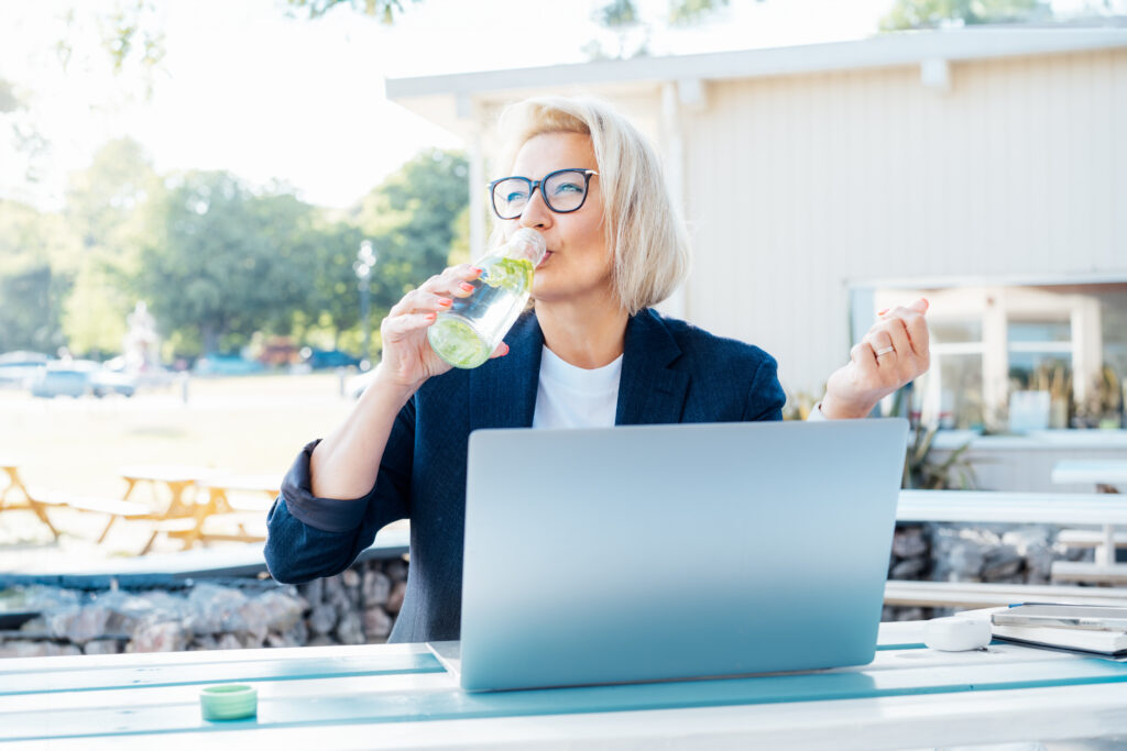A person with glasses enjoys a drink from a bottle while sitting at an outdoor table, working on a laptop. Engrossed in marketing insights, they seem to be analyzing consumer behavior. The setting, with a wooden building and trees in the background, offers inspiration amid nature's tranquility.