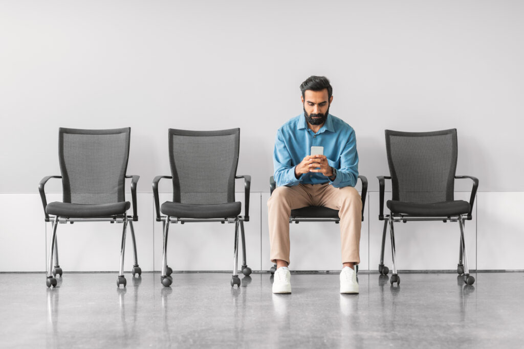 In a waiting room, a man in a blue shirt and beige pants sits on a reflective floor, using his smartphone. Surrounded by empty chairs against a plain wall, he unknowingly becomes an emblem of behavioral science in everyday settings.