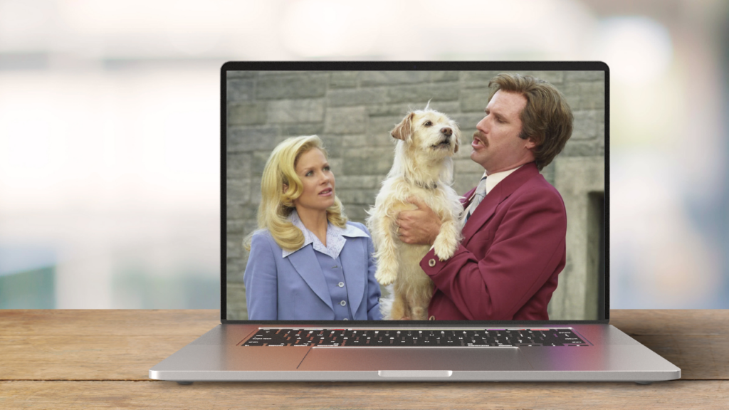 A laptop on a wooden table shows a scene where consumer behavior meets creativity: a man in a red suit holding a small dog stands beside a woman in blue, against an outdoor brick wall background.