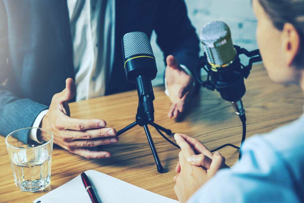 Two people sit at a table with microphones, engaged in conversation. One gestures animatedly while the other listens attentively. A notepad, pen, and glass of water adorn the table, hinting at a podcast centered on marketing psychology or behavioral science insights.