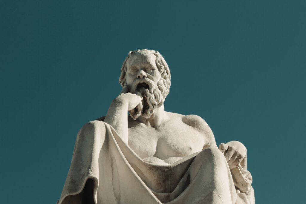 A marble statue of a seated figure with a beard, depicted in a thoughtful pose with one hand resting on their chin, resembles the essence of behavioral economics. Set against a clear blue sky, it captures the classic representation of philosophical contemplation.