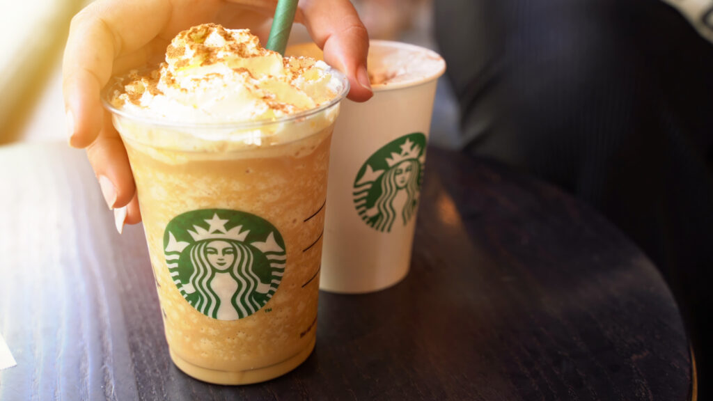 A hand holding an iced coffee, highlighted by marketing appeal, features whipped cream in a clear cup with a straw. Beside it, on the dark table, rests a white cup adorned with a distinctive green logo—perhaps influenced by insights from behavioral economics.