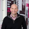A bald man wearing a dark sweater over a pink shirt smiles while standing in a modern office space, reflecting the principles of behavioral science with its walls adorned with text and images.