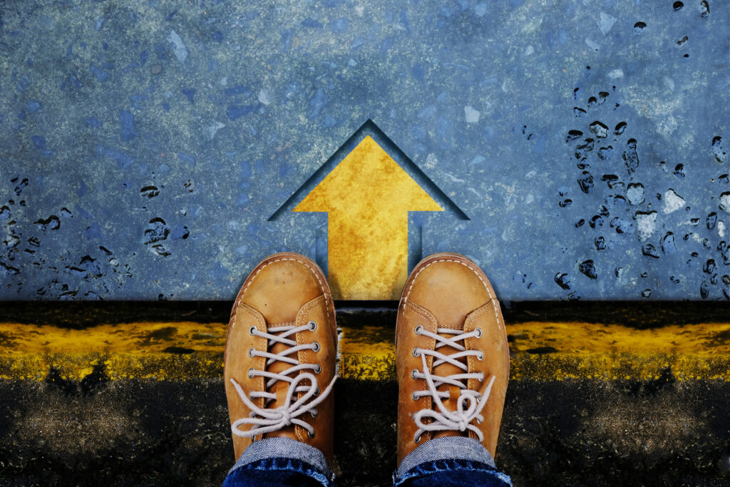 A pair of brown leather shoes standing at the edge of a pavement, facing a yellow arrow painted on the ground, pointing forward. The pavement has dark marks suggesting rainwater or wetness.