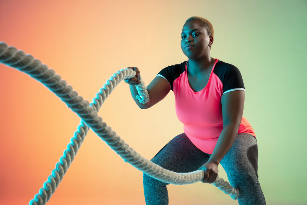 A person exercising with battle ropes against a colorful background. They are wearing a pink top and gray leggings, and appear focused and determined.