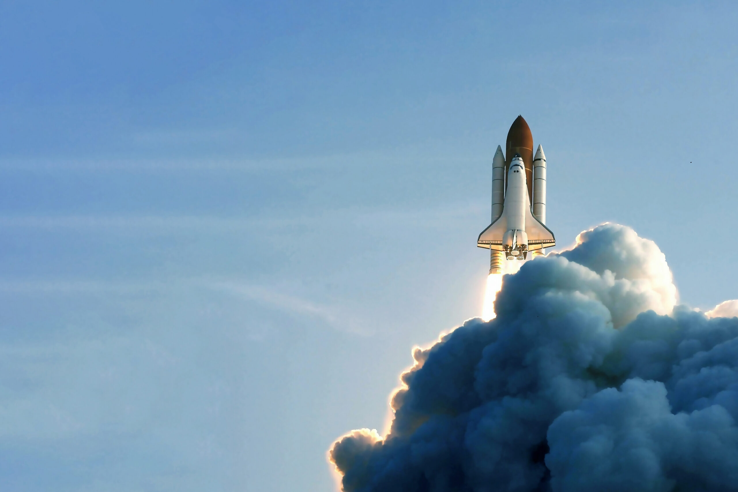A space shuttle launching into a clear blue sky, leaving behind a large plume of white smoke. Sunlight highlights the edges of the clouds, creating a dramatic and dynamic scene.