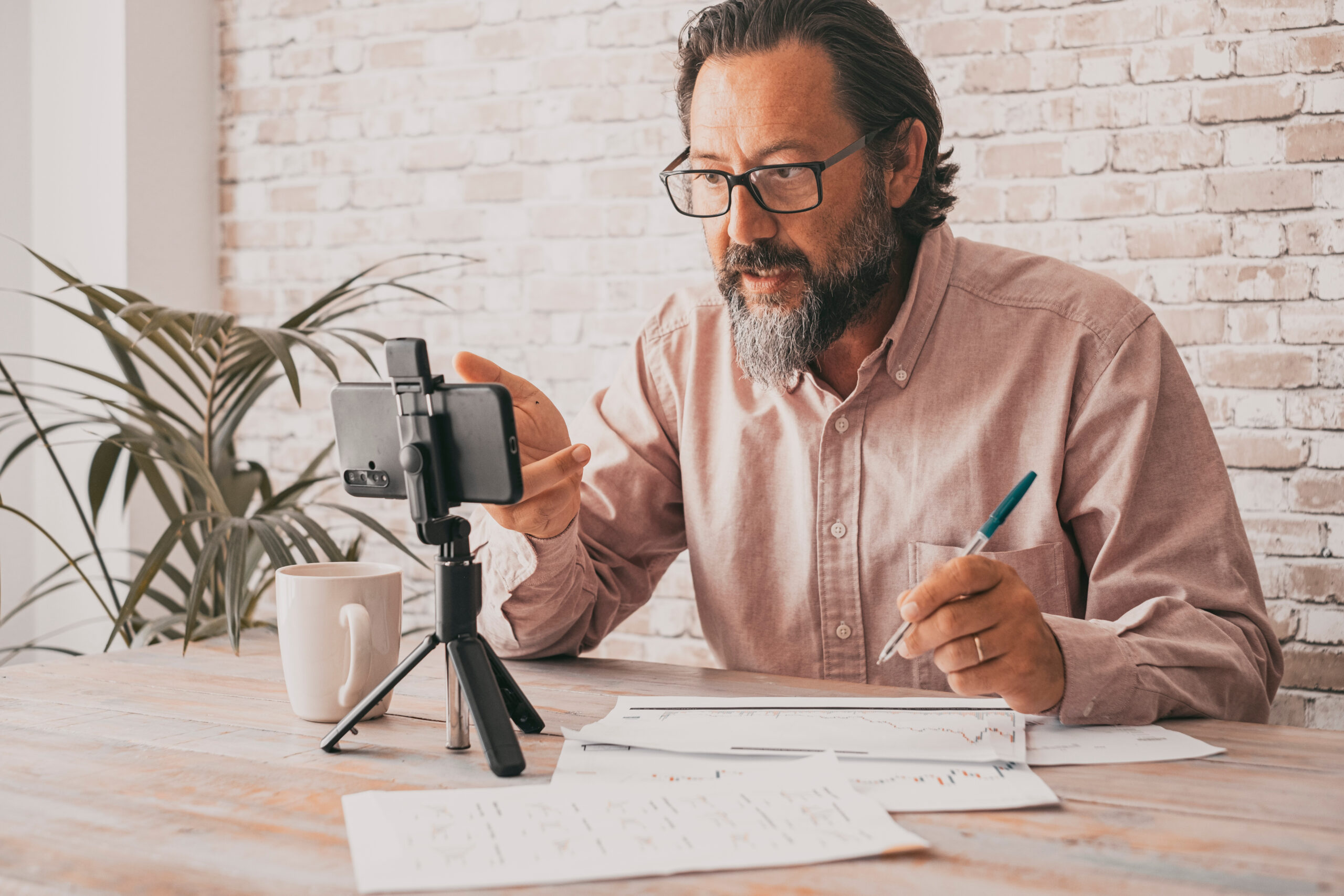 A man with glasses and a beard sits at a table with papers, a pen, and a smartphone on a tripod. He appears focused, gesturing towards the phone. A white mug is nearby, and a potted plant is in the background.