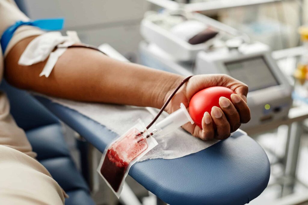 A person donating blood, with an arm resting on a blue cushion. A tube is connected to a blood collection bag. The person is squeezing a red stress ball to aid blood flow. Medical equipment is visible in the background.