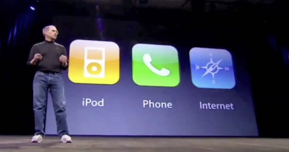 A person in a black turtleneck and jeans is on stage speaking. Behind them, a large screen displays three icons labeled "iPod," "Phone," and "Internet.