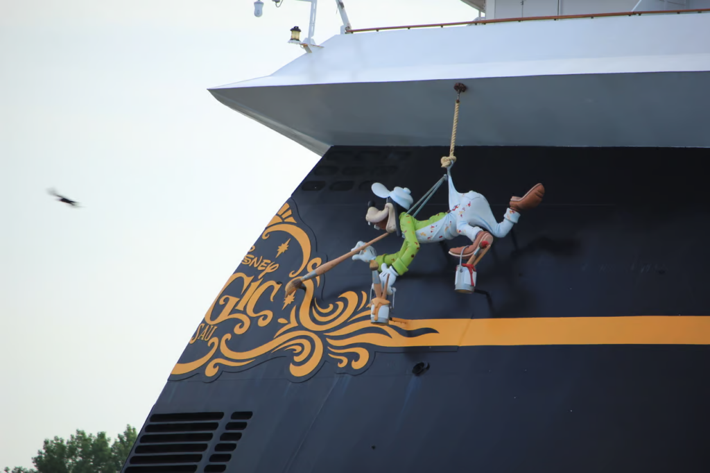 A cartoon character in a green outfit is hanging from a rope on the side of a large ship, appearing to paint a decorative pattern. The ship is black with orange and gold accents. A small flying insect is nearby.
